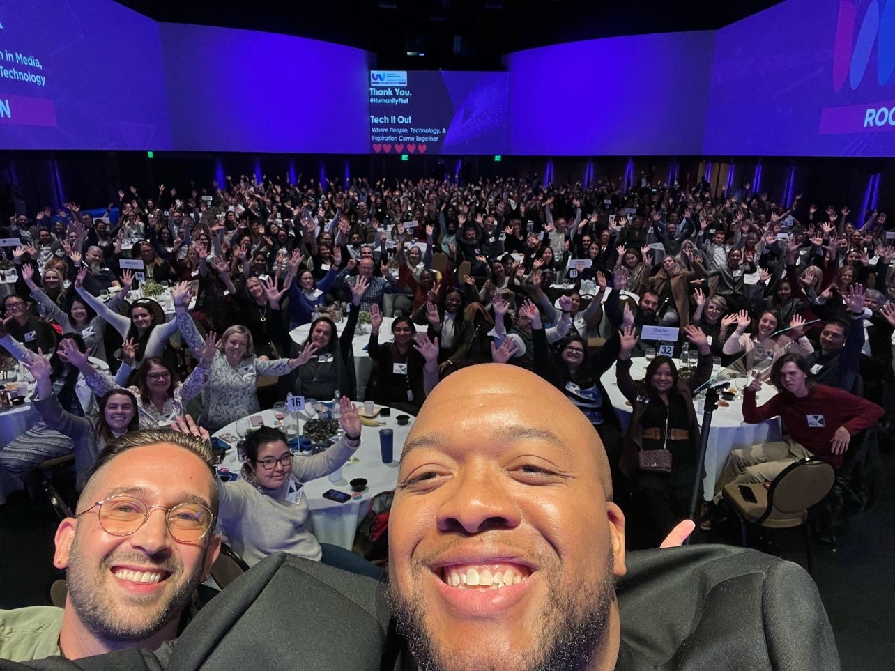 Christopher Lafayette and a fellow speaker smiling in front of a large audience raising their hands at Tech It Out.