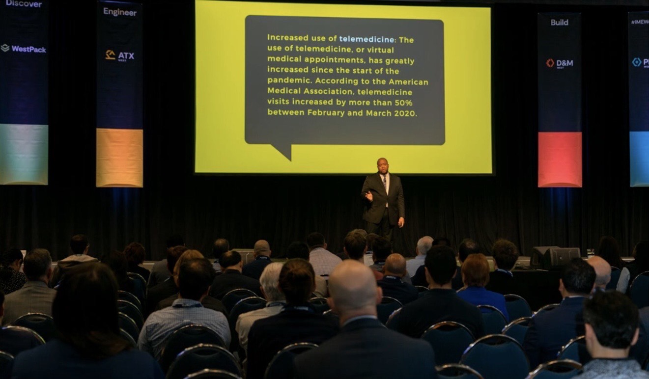 Christopher Lafayette speaking beneath a large presentation screen, with a seated audience in the foreground.