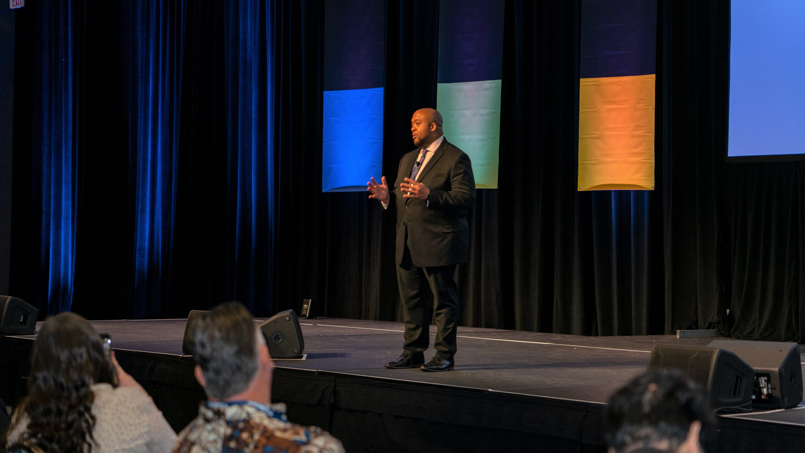 Christopher Lafayette gesturing on stage between blue and amber panels.