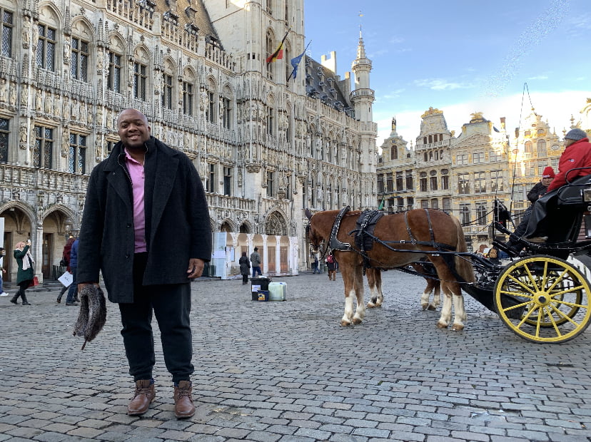 Christopher Lafayette standing in a cobblestone square with ornate European buildings and a horse-drawn carriage.
