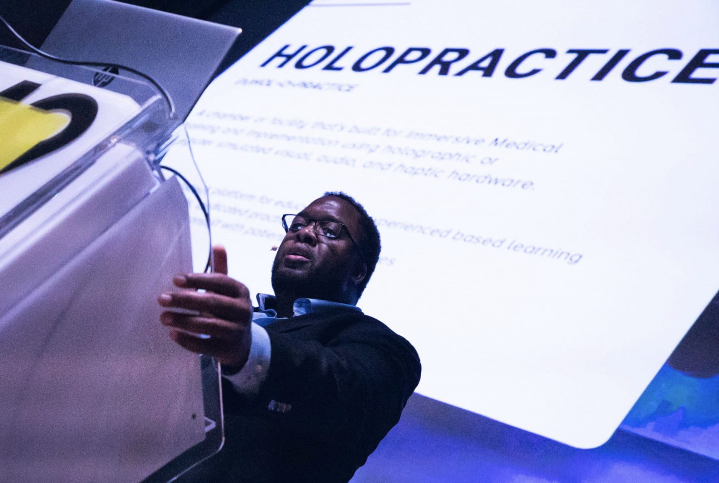 Christopher Lafayette speaks beside a lectern with Holopractice projected behind him.