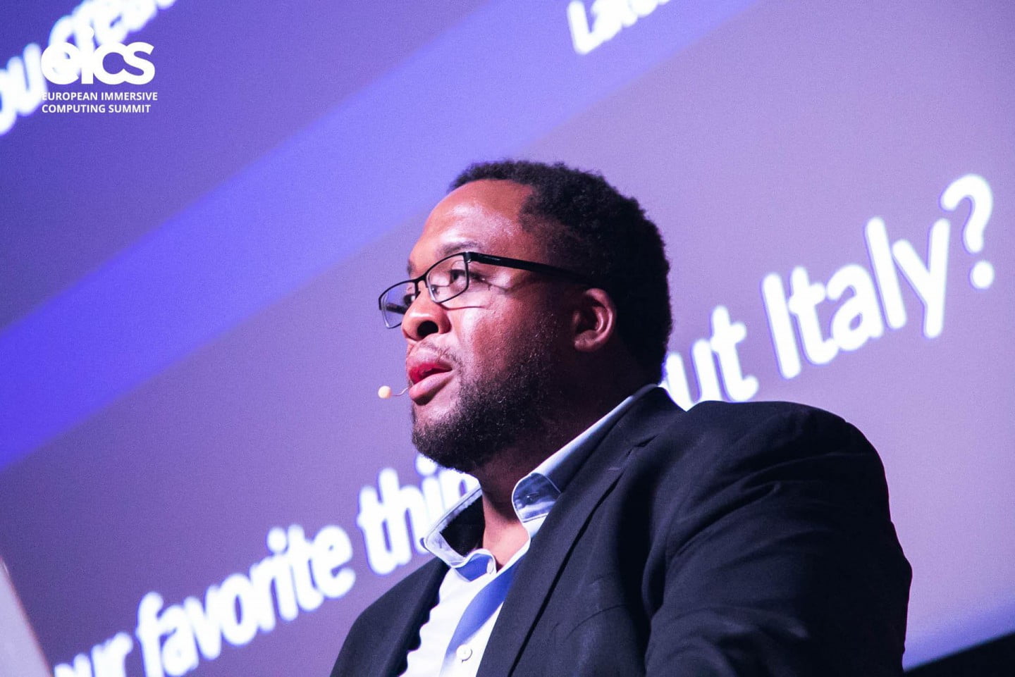 Close-up of Christopher Lafayette speaking on stage, with the EICS logo and purple projected screen behind him.