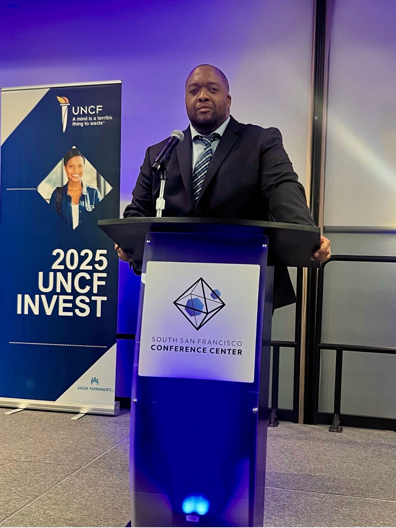Speaker at a lectern beside a 2025 UNCF INVEST banner at the South San Francisco Conference Center.