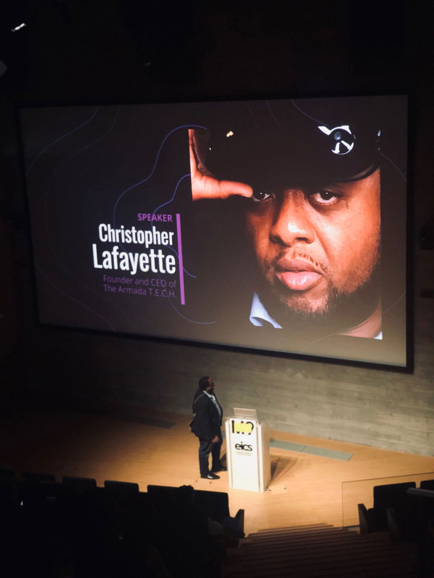 Christopher Lafayette stands beside an EICS lectern beneath a large projected speaker portrait and name.