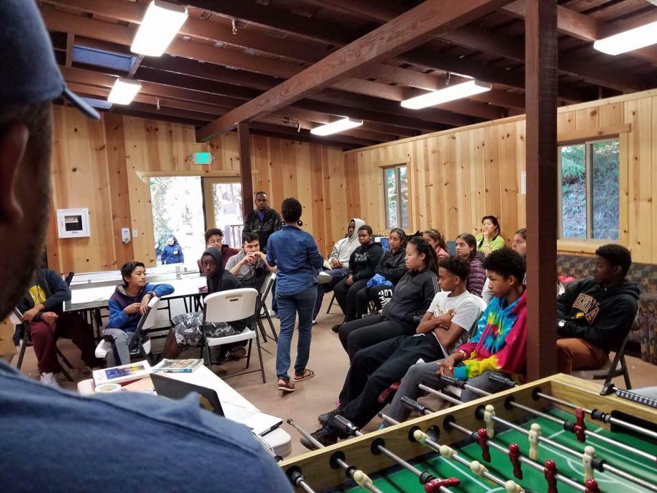 Young people and adults gather for a discussion in a wood-paneled room, with a foosball table in the foreground.